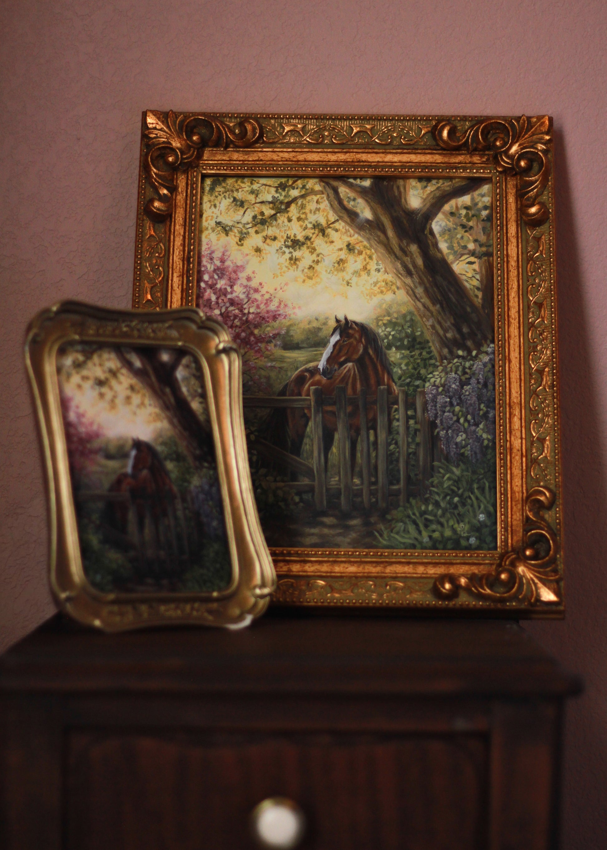 Two framed paintings of horses in a garden setting on a wooden surface.
