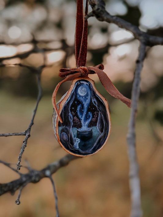 A teardrop-shaped wood ornament hanging from a brown ribbon, featuring a painted celestial scene with a silver moon, blue and white clouds, and a dark night sky, set against a blurred outdoor background of tree branches.